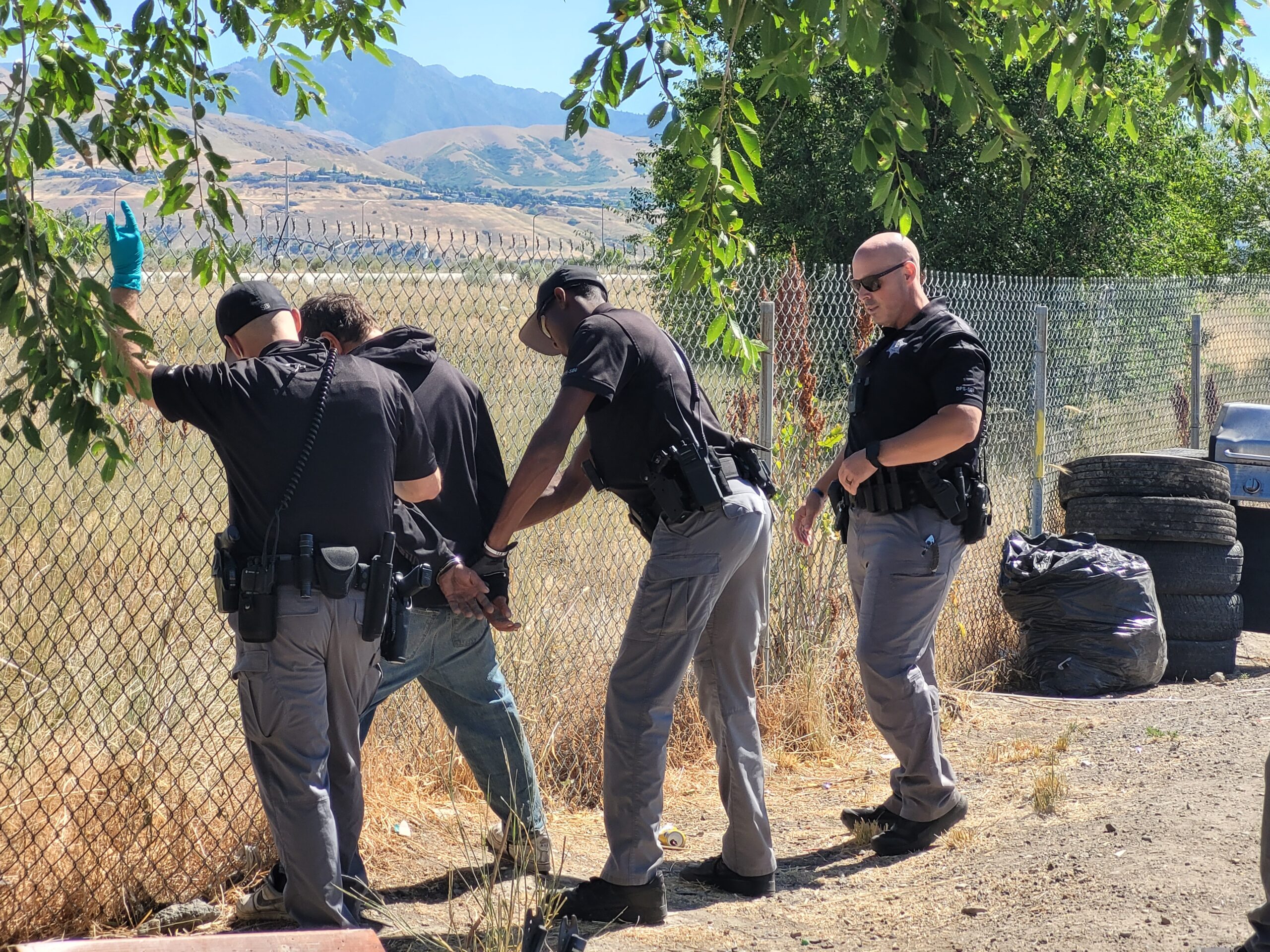 Three members of SBI are handcuffing a person who is standing up against a chain link fence
